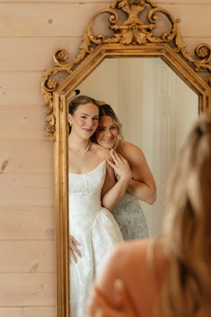 Two brides share a tender moment reflected in an ornate gold mirror, one in a white wedding gown with pearl necklace.