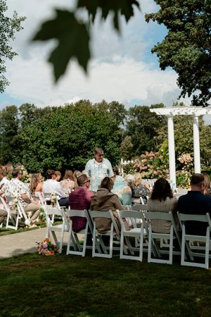 Guests seated in white chairs at an outdoor wedding ceremony near a white arbor surrounded by lush greenery.