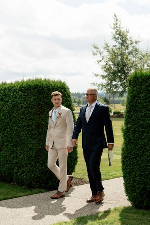 Two men in suits walk along a garden path flanked by tall hedges at an outdoor wedding ceremony.