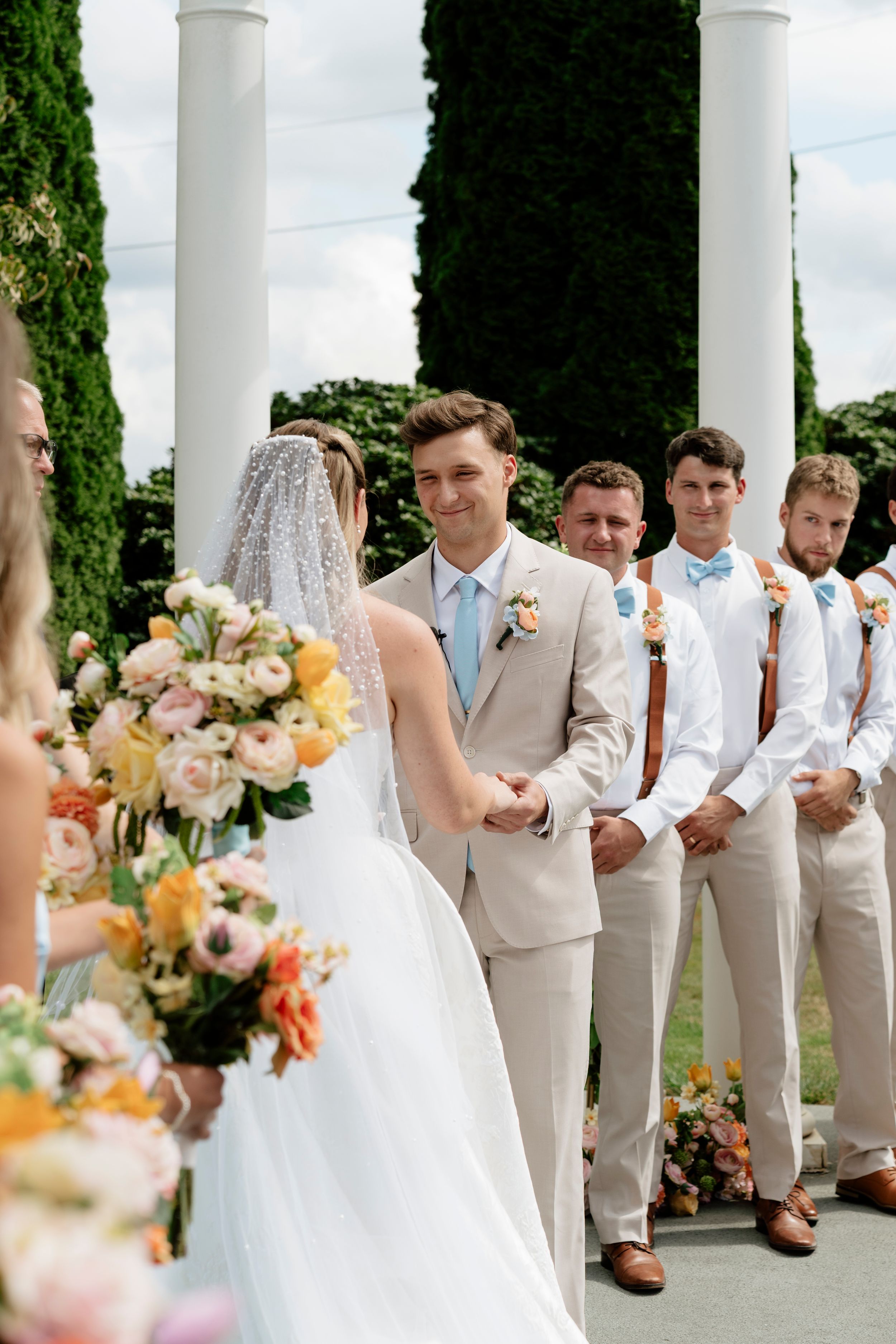 Groom in beige suit smiles at bride during outdoor wedding ceremony with groomsmen in suspenders standing nearby.