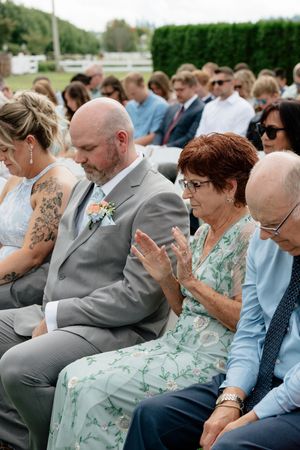 Wedding guests seated outdoors, clapping during ceremony, wearing formal attire including gray suit and floral dress.