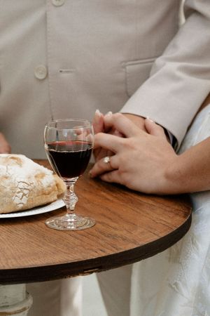 Couple holding hands at a wooden table with red wine and bread, wearing wedding rings.