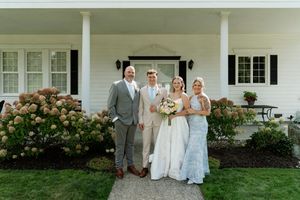 Wedding party of four posing in front of a white farmhouse with blooming hydrangeas on a sunny day.