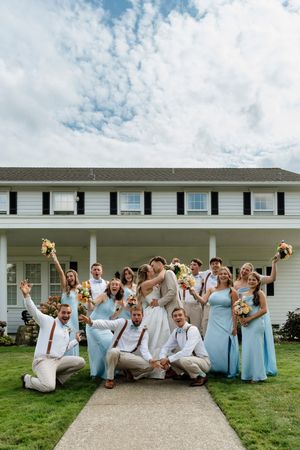 Wedding party celebrating outside a white colonial home, bridesmaids in light blue dresses with groomsmen in tan suits.