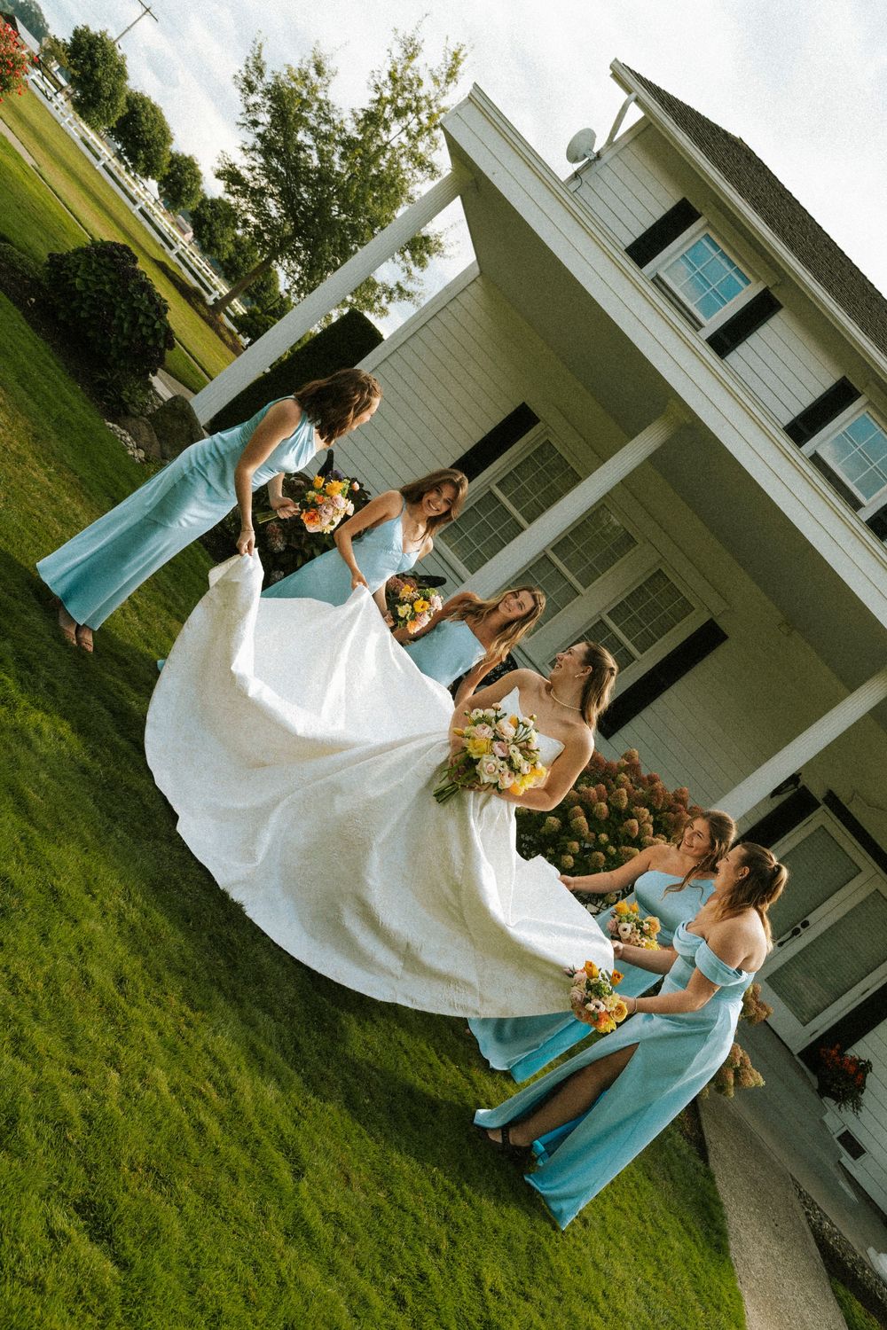 Bride in white gown with bridesmaids in light blue dresses holding bouquets on green lawn outside white house.