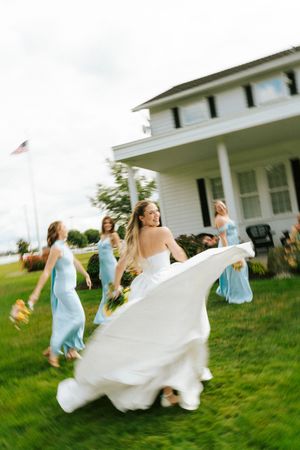 Smiling bride in white gown twirling with bridesmaids in light blue dresses running across green lawn.