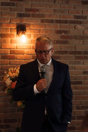 Man in navy suit speaking into microphone at wedding reception against rustic brick wall backdrop.