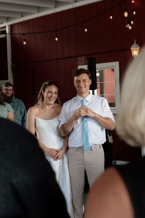 Smiling bride and groom stand together at their wedding reception in a rustic red barn venue with string lights.