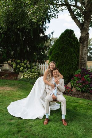 Bride in white gown embracing groom in tan suit seated in chair surrounded by lush garden greenery.