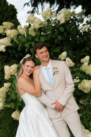Bride and groom posing together in front of blooming white hydrangea bushes, groom in tan suit with blue tie.
