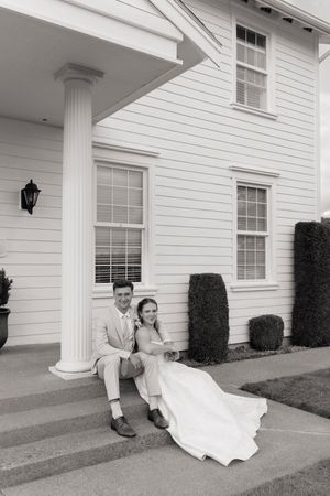 Black and white photo of bride and groom sitting on steps of elegant white colonial-style wedding venue.