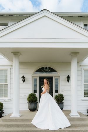 Bride in flowing white ball gown twirling in front of a grand white colonial mansion with tall columns.
