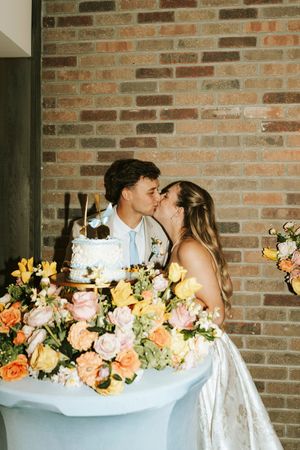 Newlyweds share a kiss at their wedding cake table adorned with colorful peach and yellow floral arrangements.