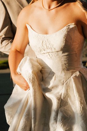 Close-up of a strapless ivory wedding gown with textured fabric and pearl necklace in golden sunlight.