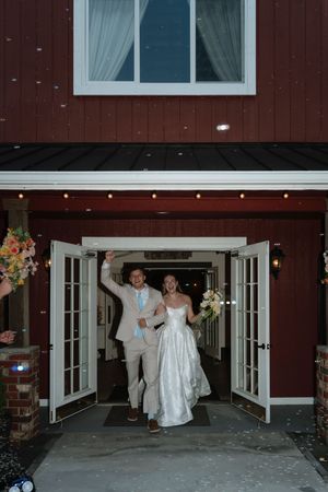 Joyful newlyweds exit a red barn wedding venue through open French doors, bride in white gown holding bouquet, groom in tan suit.