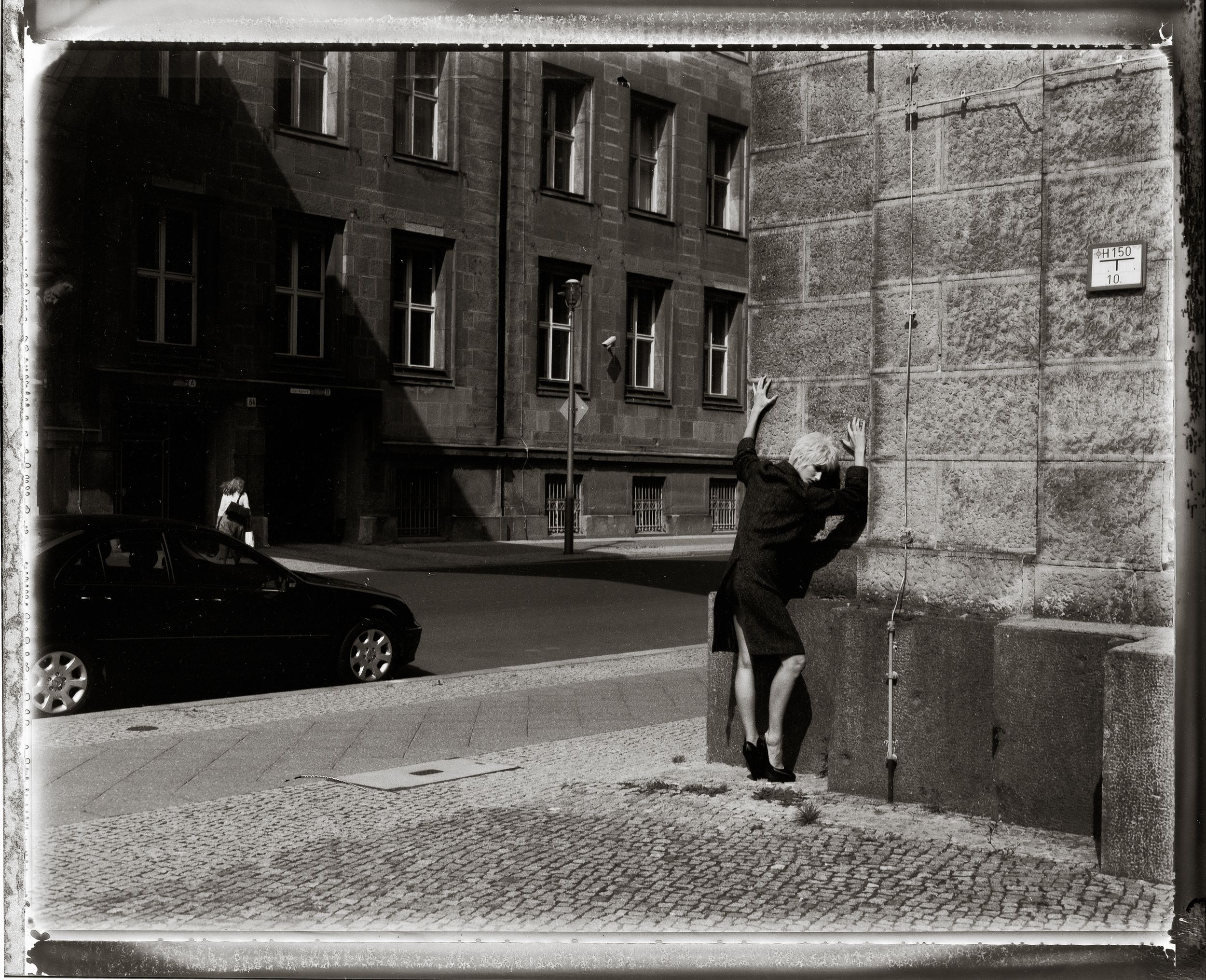Black and white photo of a person leaning against a stone building corner on a cobblestone street in a European city.