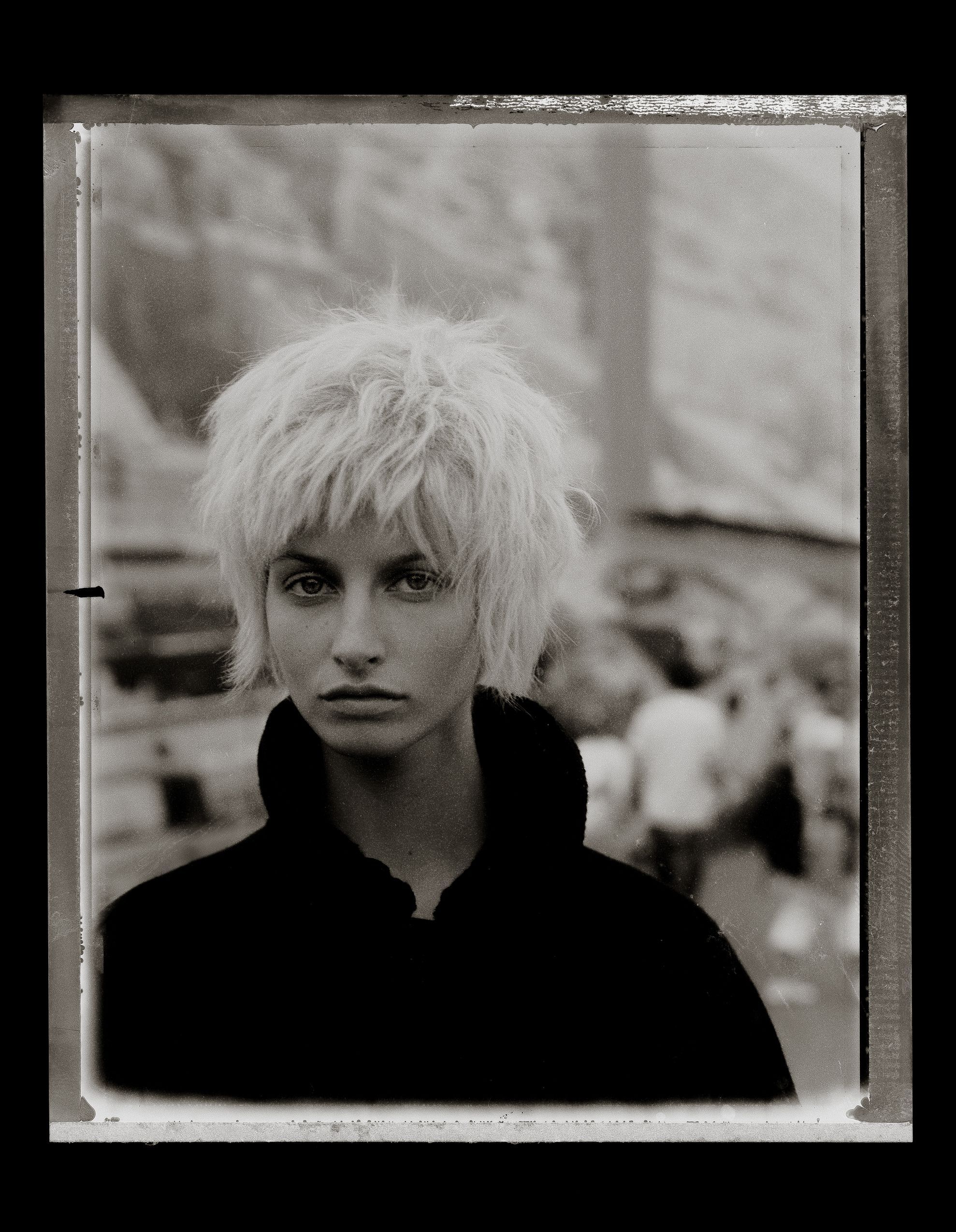 Black and white portrait of a young woman with short blonde messy hair wearing a dark jacket outdoors.