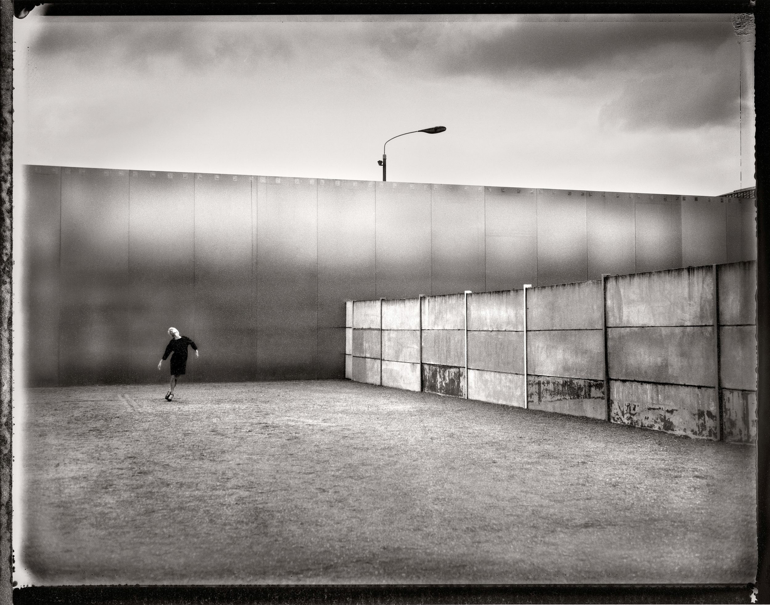 Black and white photo of a lone figure walking near a large concrete wall with a street lamp overhead.