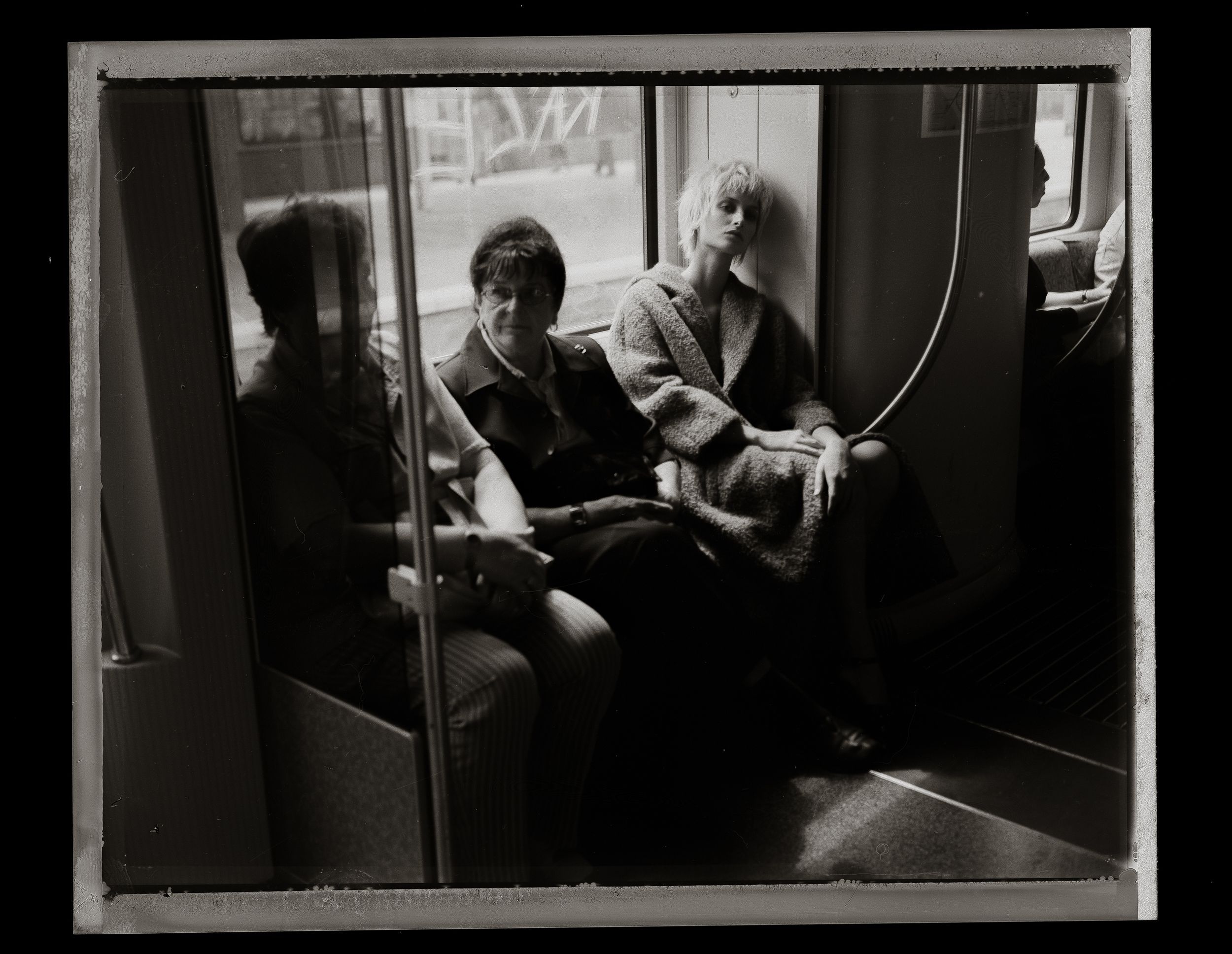 Black and white photo of two people sitting on a subway train, with a reflection visible in the window beside them.