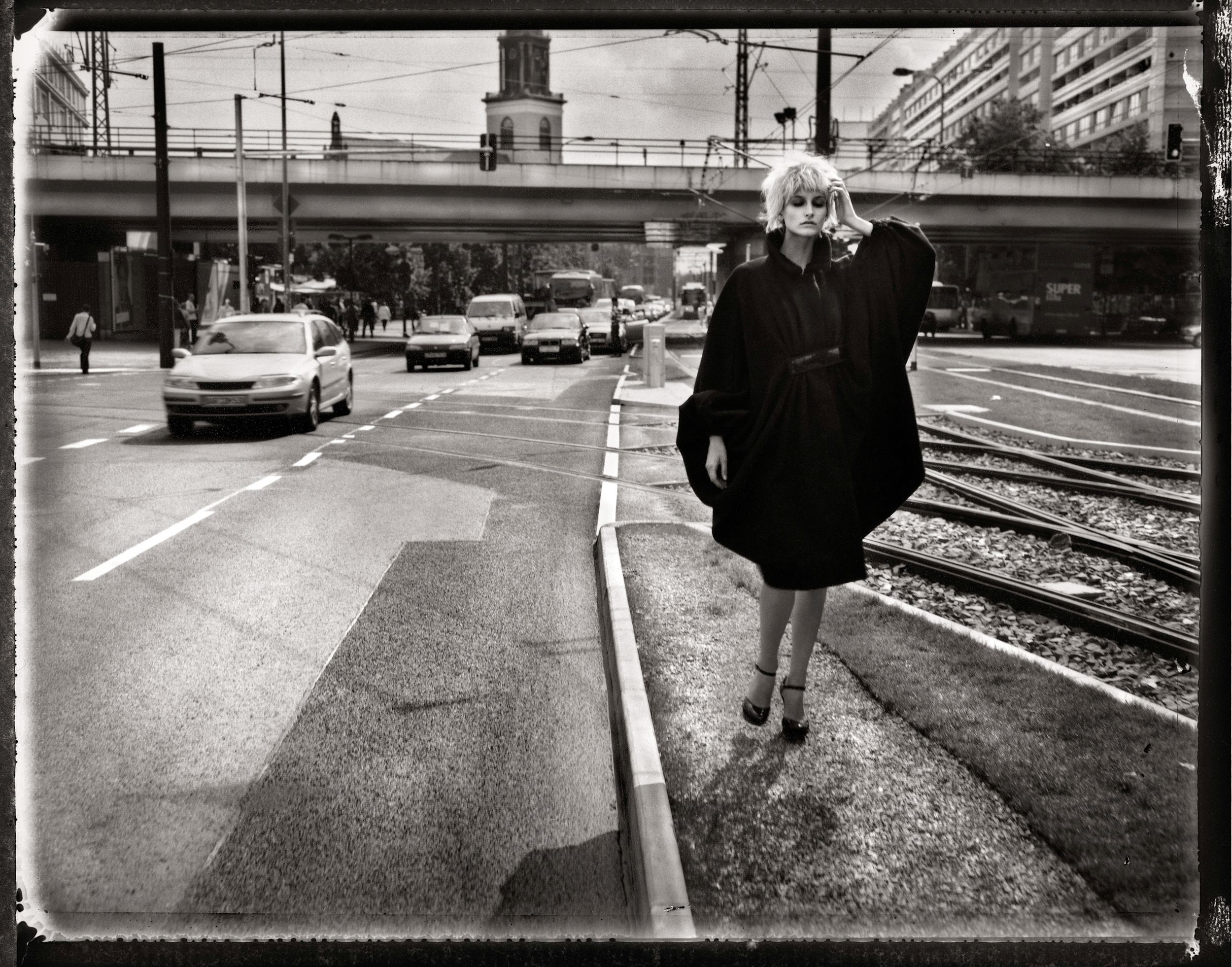 Black and white photo of a blonde woman in a black coat standing on a city street with traffic and a bridge.