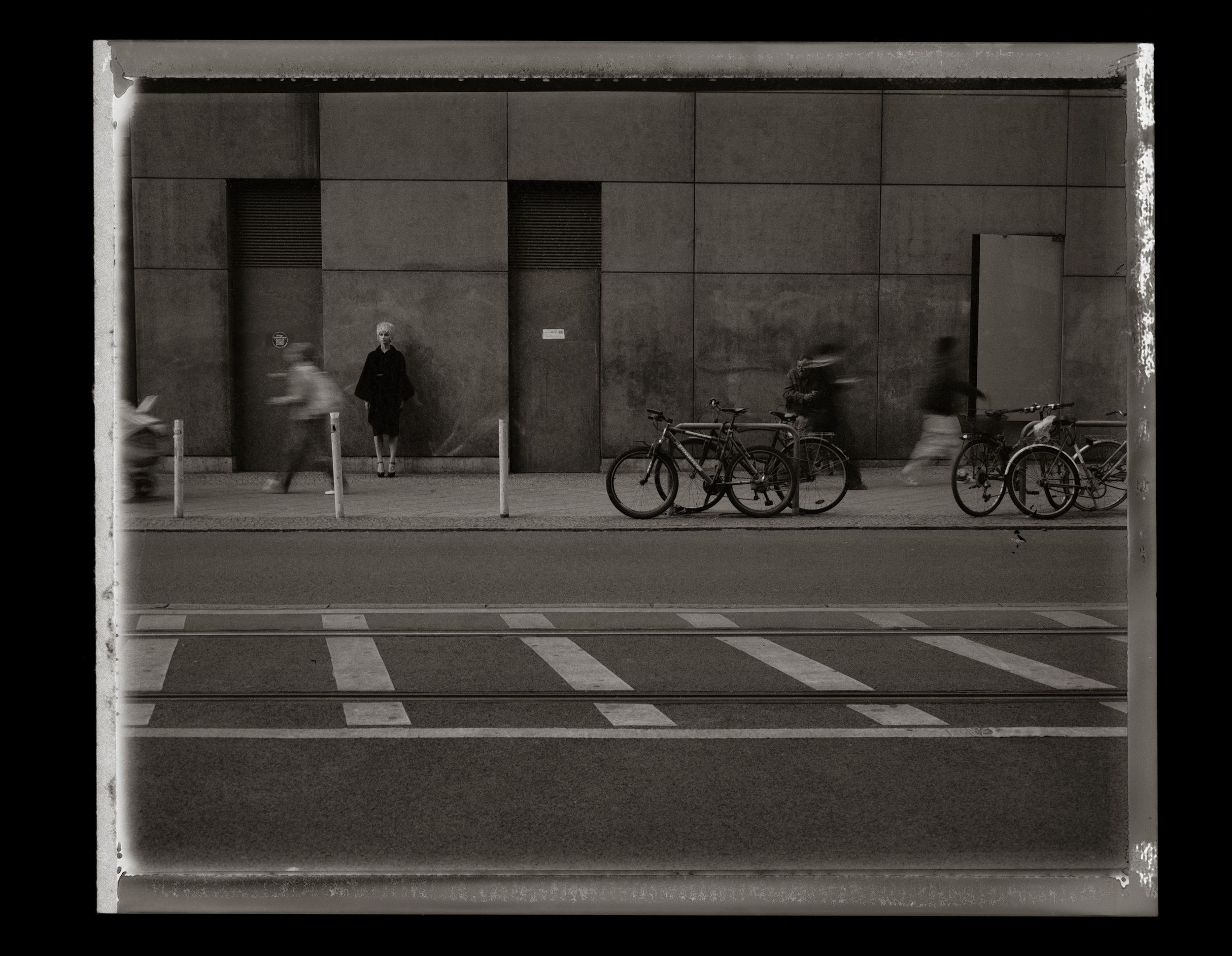 Black and white photo of blurred pedestrians and parked bicycles on an urban sidewalk beside a concrete building.