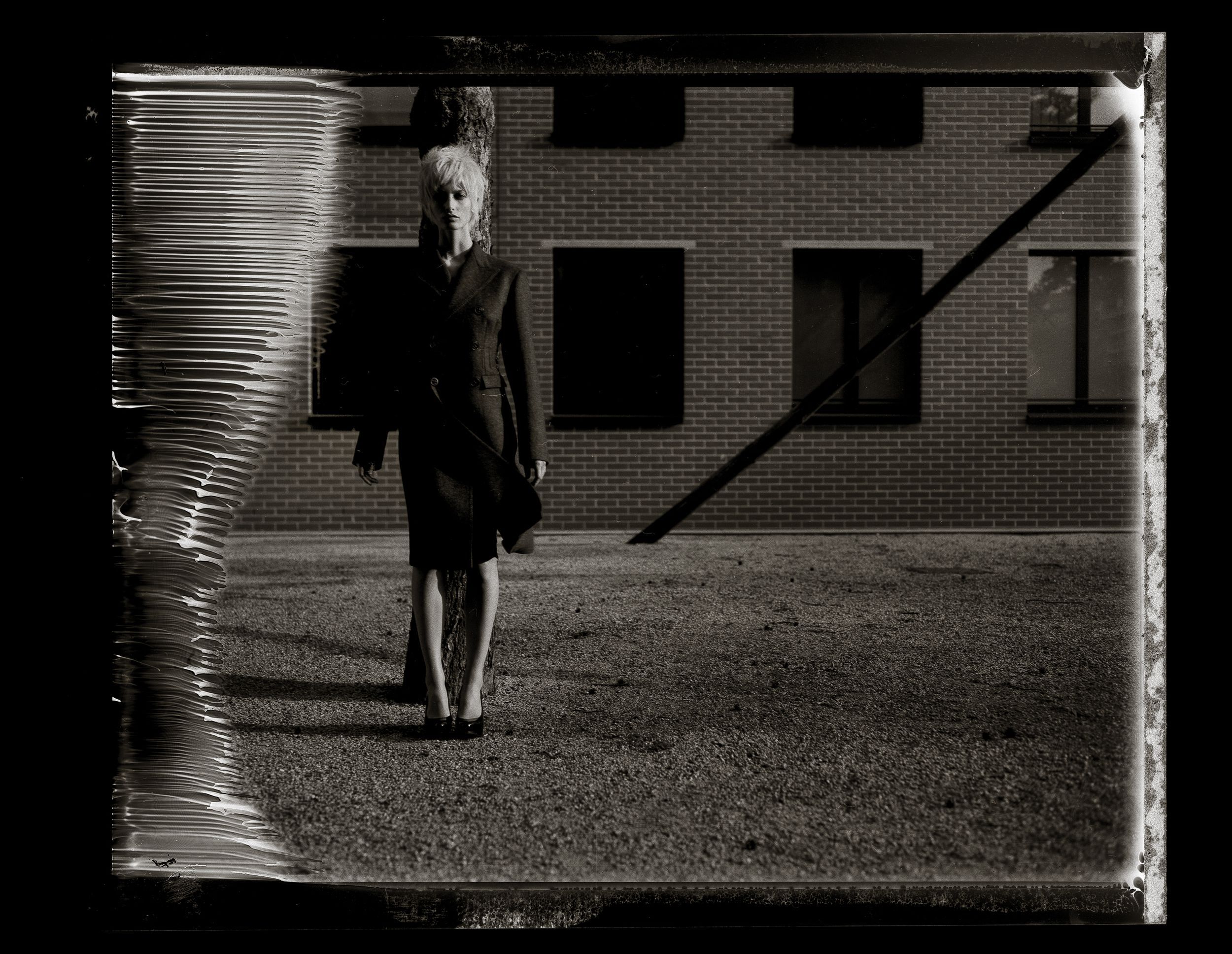 Vintage black and white photo of a woman in dark clothing standing outside a brick building with dramatic shadows.