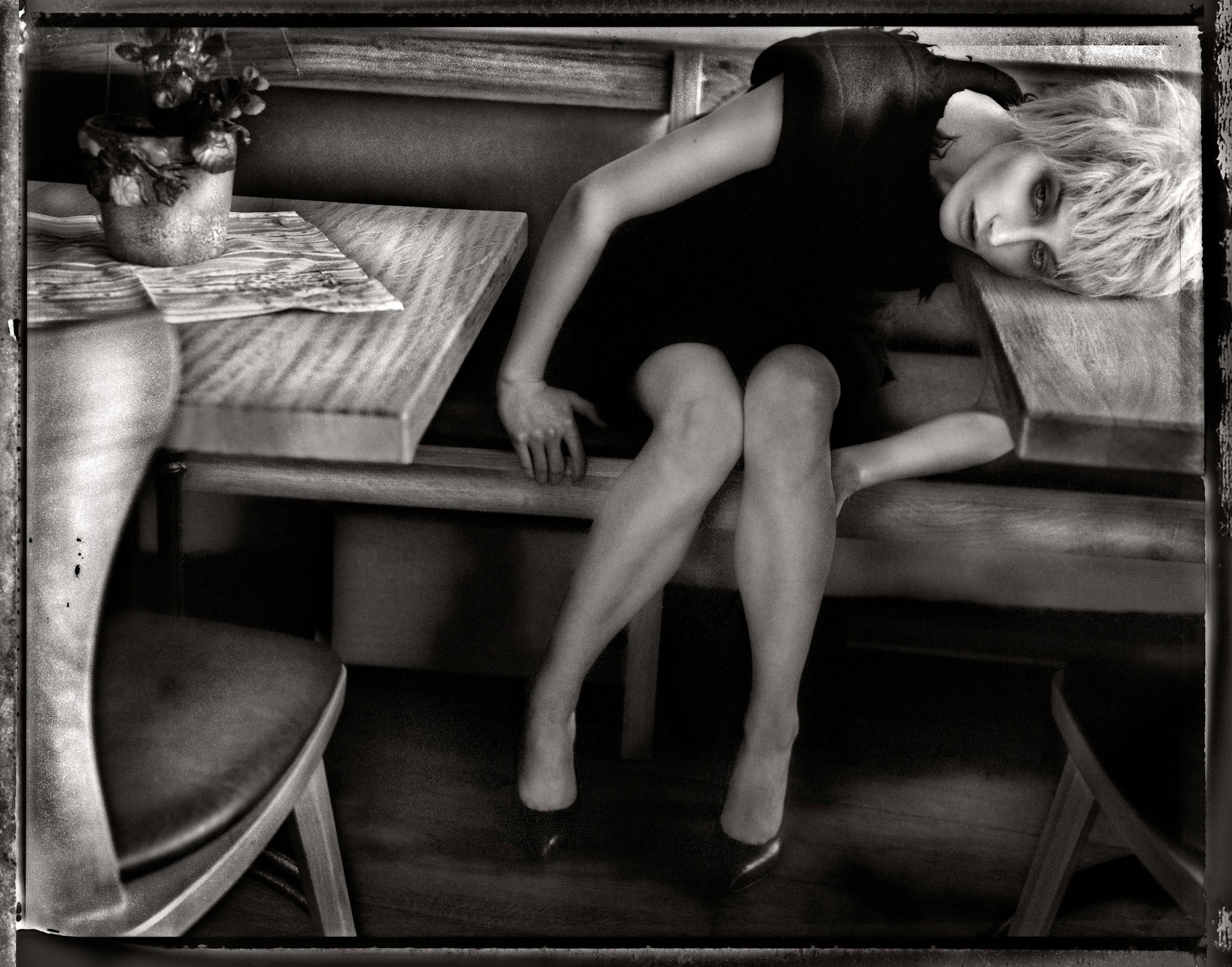 Black and white photo of a woman in a black dress and heels reclining on a wooden bench in a diner.