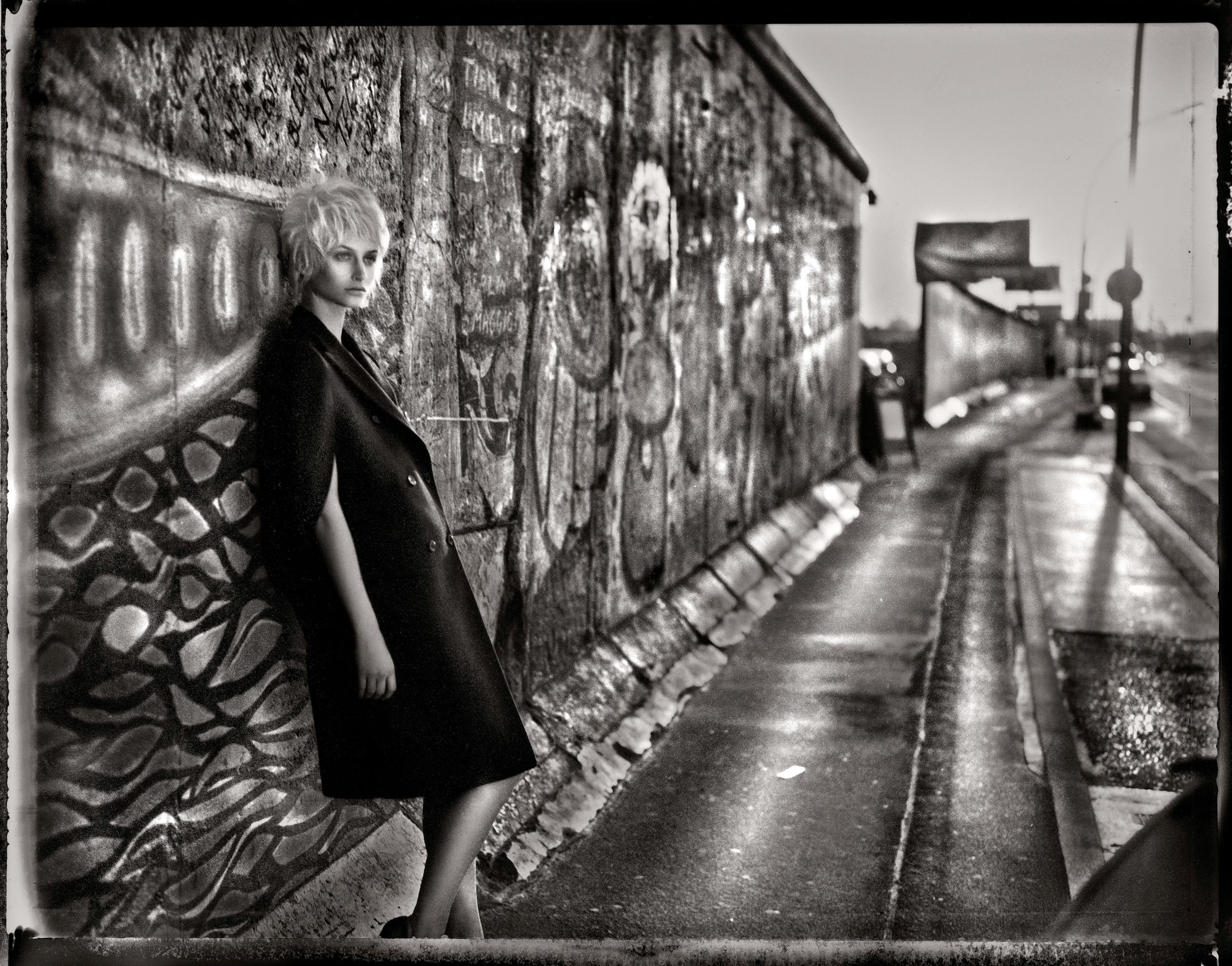 Black and white photo of a woman in dark coat leaning against graffiti-covered Berlin Wall on a wet sidewalk.