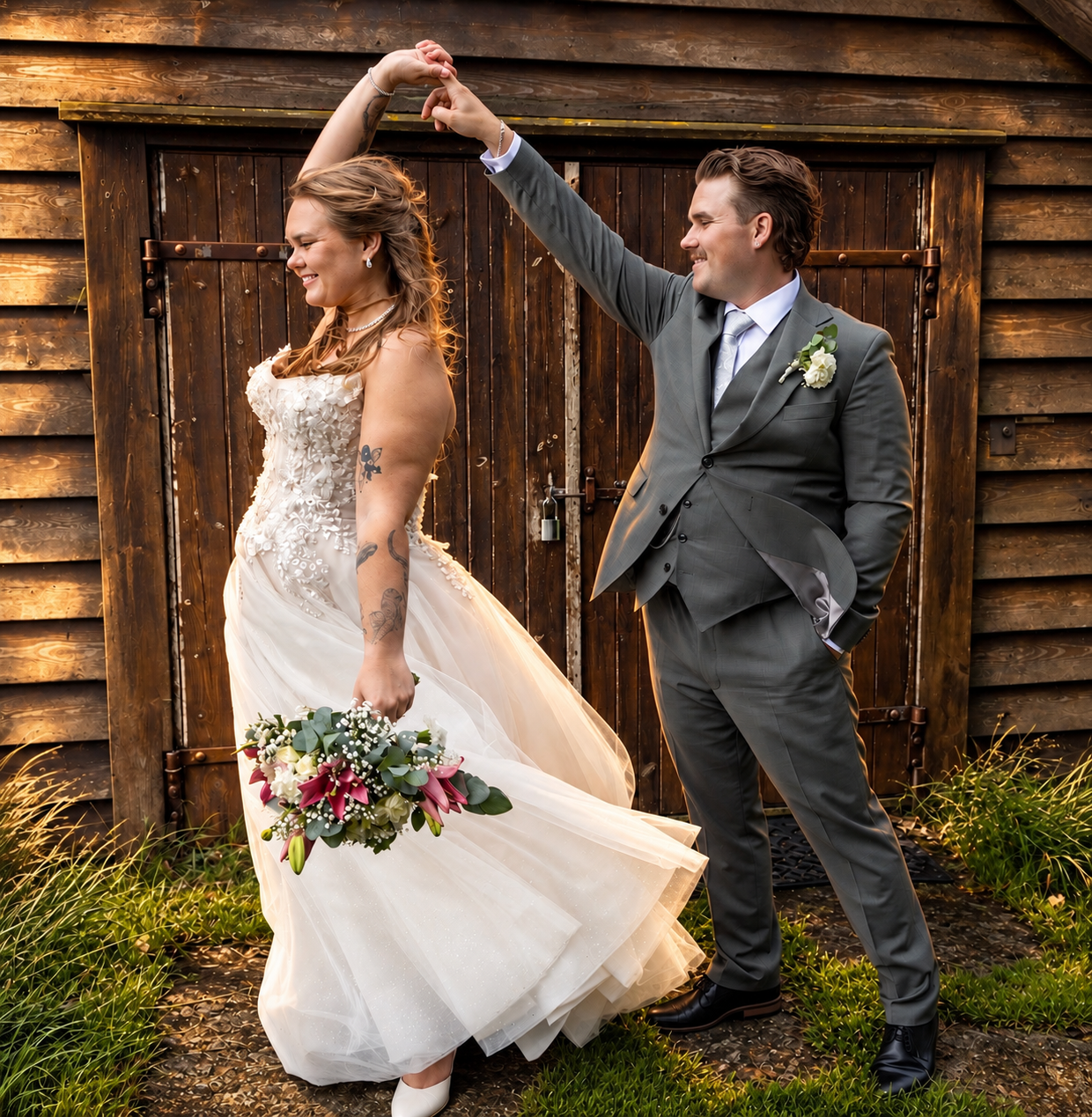 Bride and groom dancing joyfully outside rustic barn doors, bride in lace gown holding bouquet, groom in gray suit.
