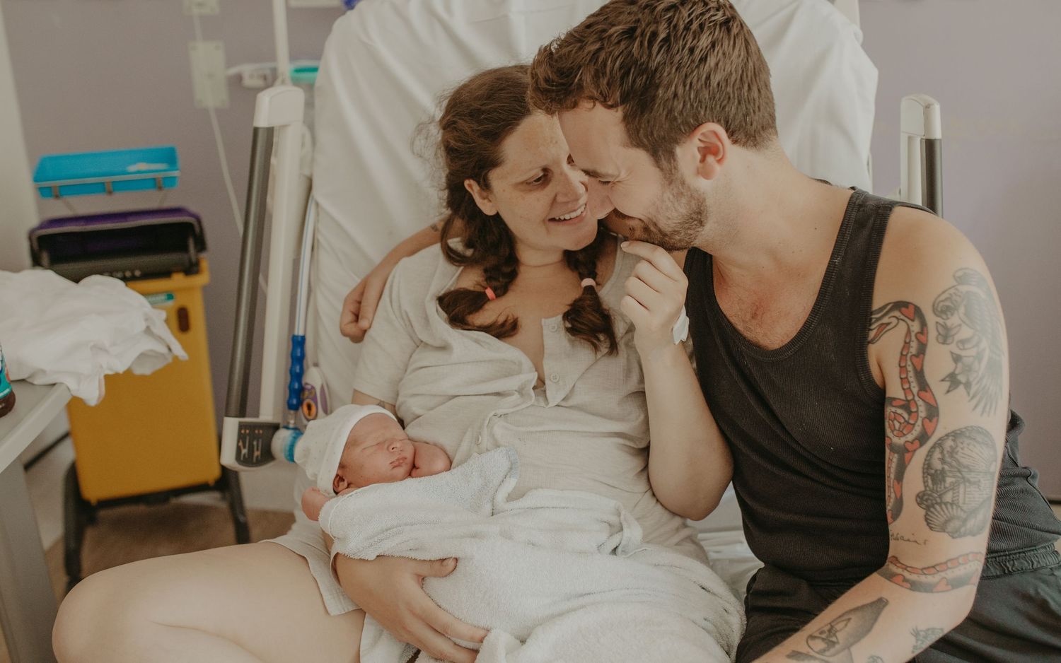 New parents share a tender moment in hospital bed, mother holding newborn baby while tattooed father kisses her hand.