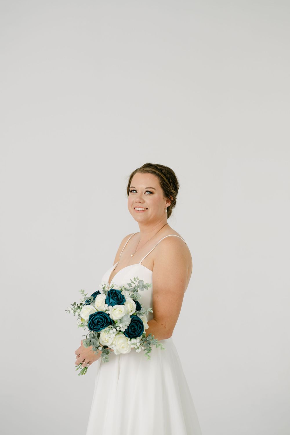Bride in white gown holding teal and white rose bouquet with greenery, smiling against light gray background.
