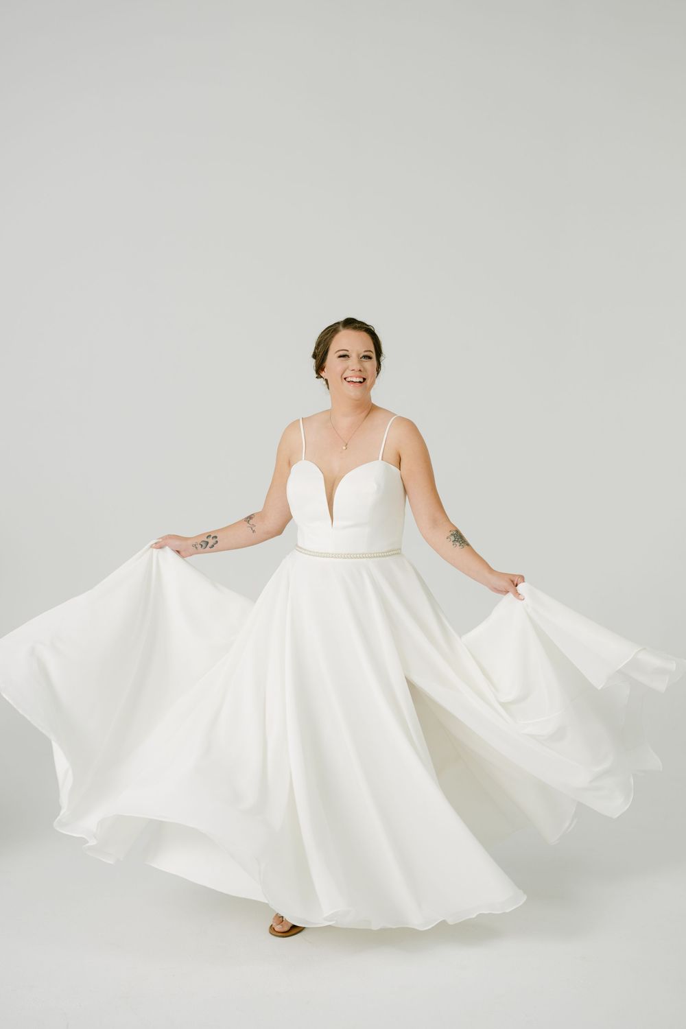 Smiling bride in white spaghetti strap wedding gown twirling skirt against white background.