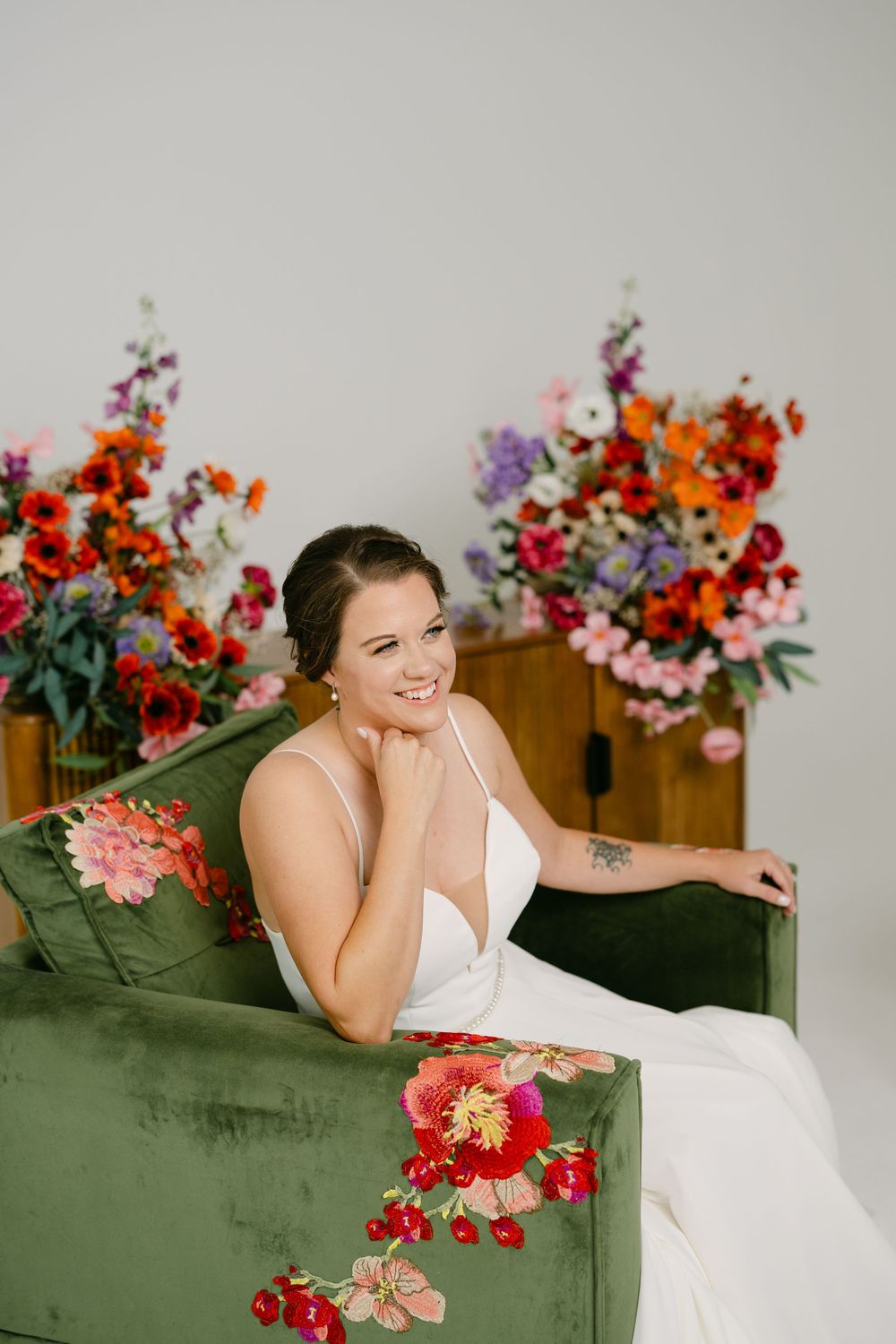 Smiling bride in white gown seated on green velvet floral sofa with colorful flower arrangements in background.