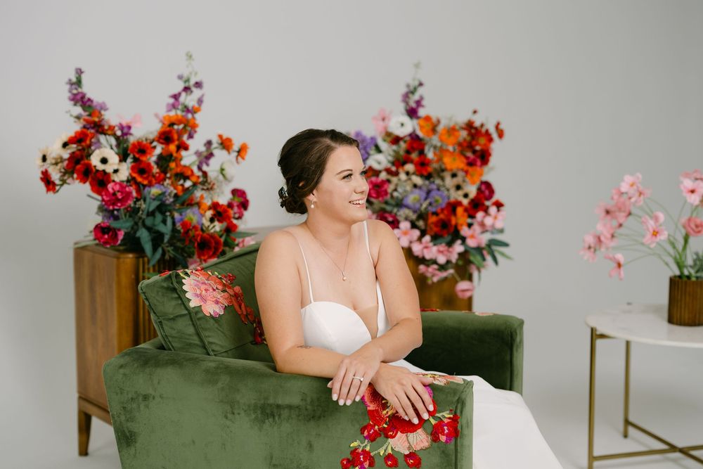 Bride in white dress sits in green velvet chair surrounded by vibrant colorful floral arrangements in bright studio.