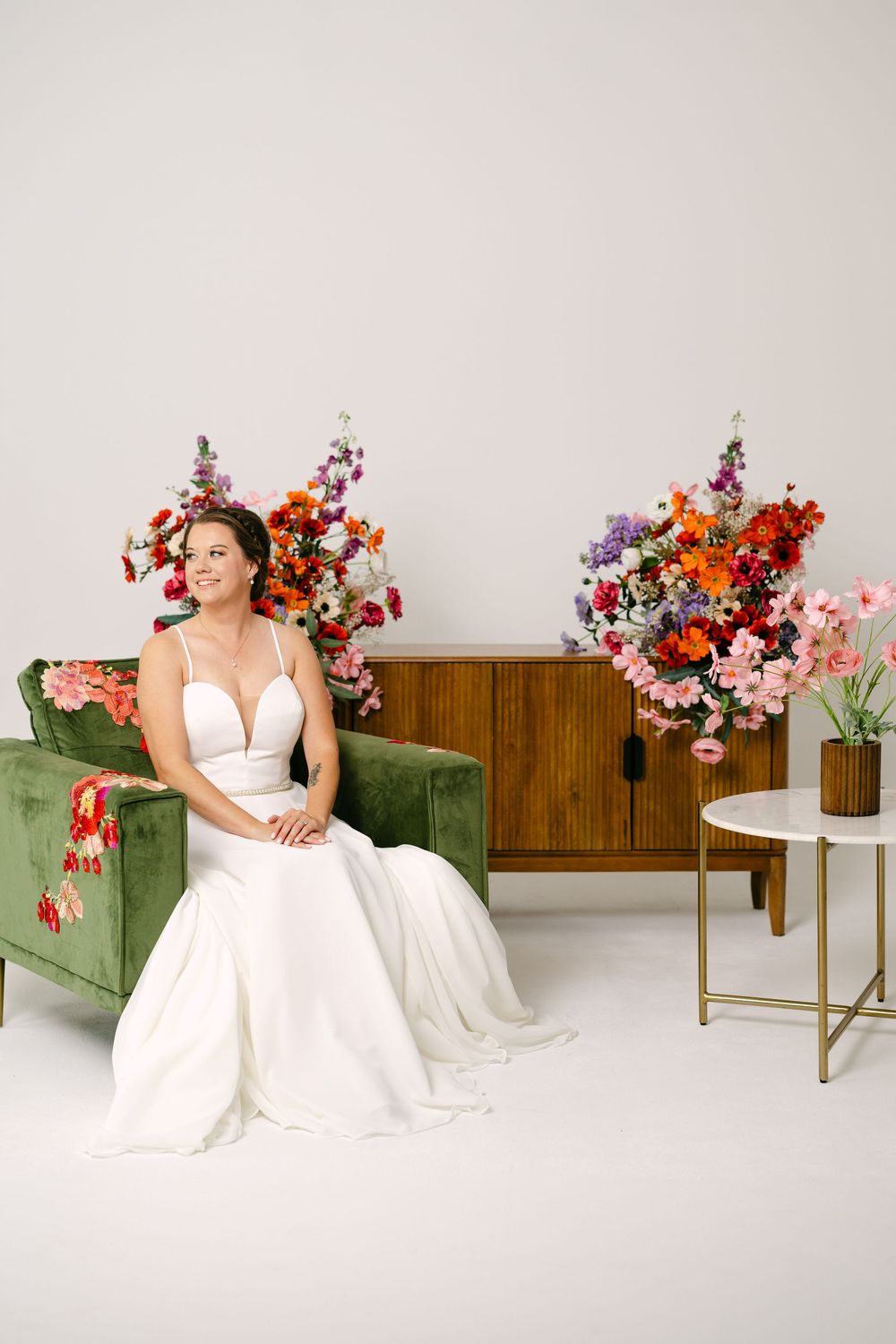Bride in white gown seated on green velvet sofa surrounded by colorful floral arrangements in a bright studio.