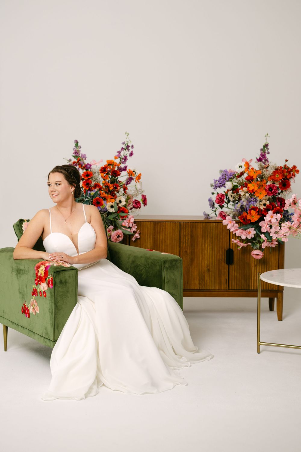 Bride in white gown seated on green velvet sofa surrounded by colorful floral arrangements against white backdrop.