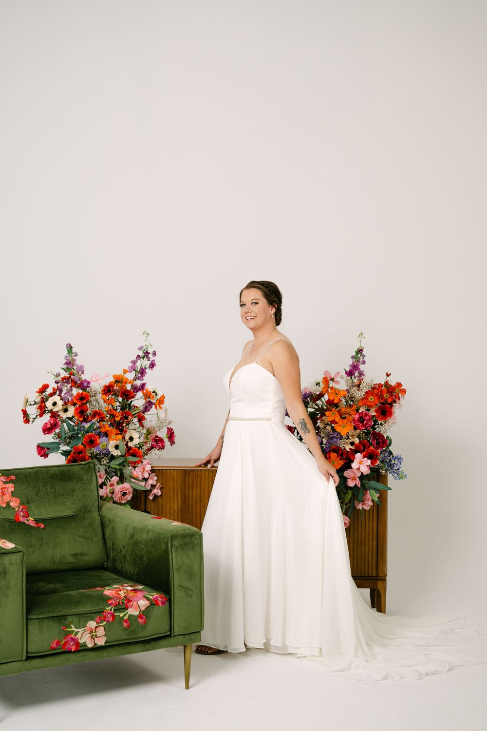 Bride in white gown poses by green velvet sofa with vibrant floral arrangements in a bright studio.