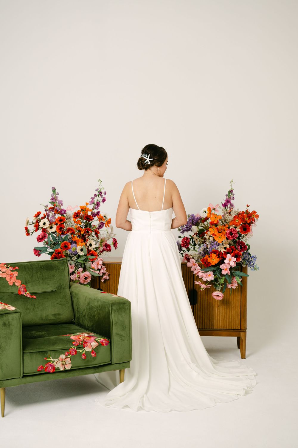 Bride in white gown with train stands between colorful floral arrangements and green velvet chair.