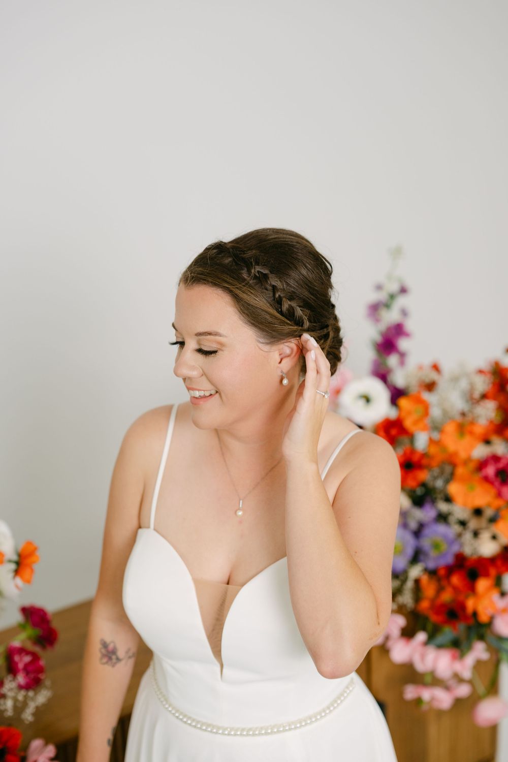 Bride in white gown adjusting earring, braided updo, floral backdrop with colorful blooms, smiling softly.