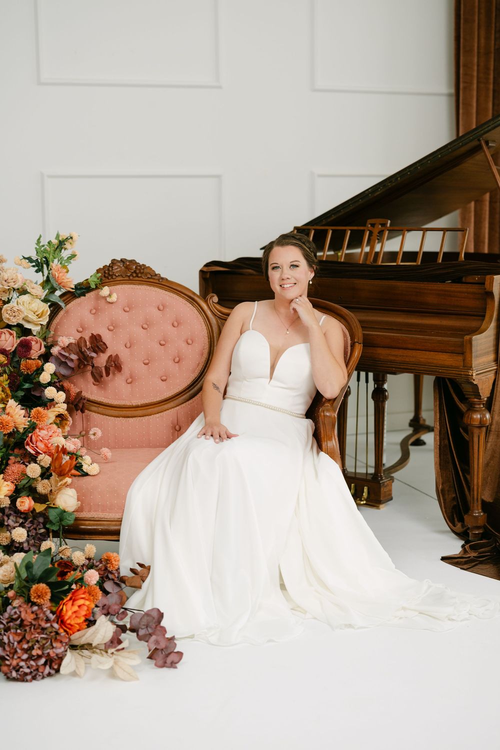 Bride in white gown seated on pink vintage chair beside grand piano and colorful floral arrangements.