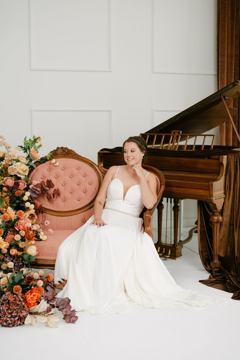 Bride in white gown sits in pink tufted chair beside grand piano and autumn floral arrangement.