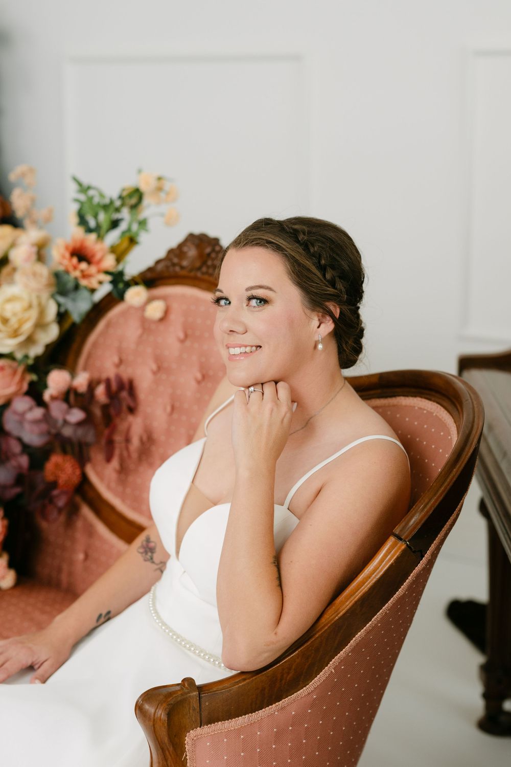 Smiling bride in white gown seated in vintage pink velvet chair with floral arrangement, wearing pearl earrings and braided updo.