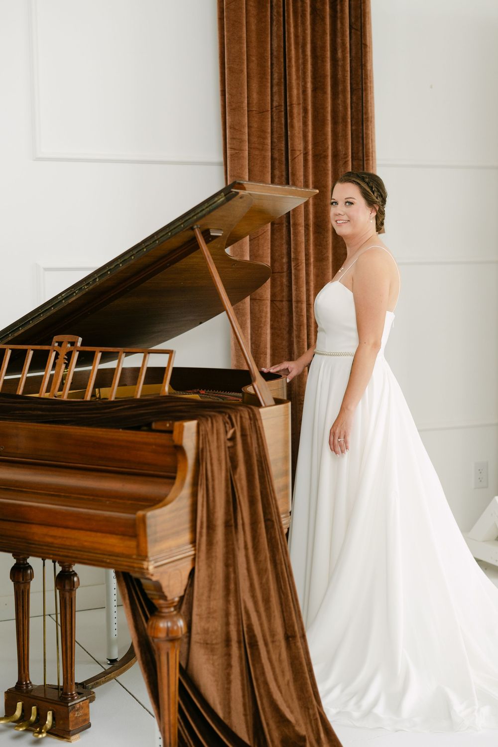 Bride in white strapless gown standing beside an elegant wooden grand piano with brown velvet drape.
