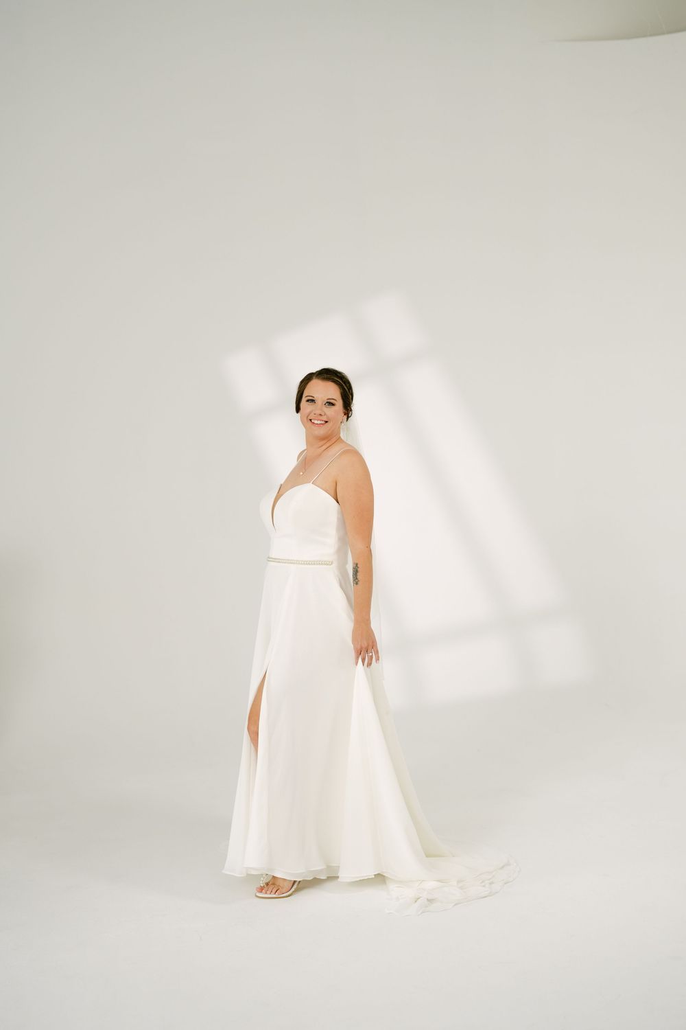 Elegant bride in a white spaghetti-strap gown with a slit standing in a sunlit white studio space.