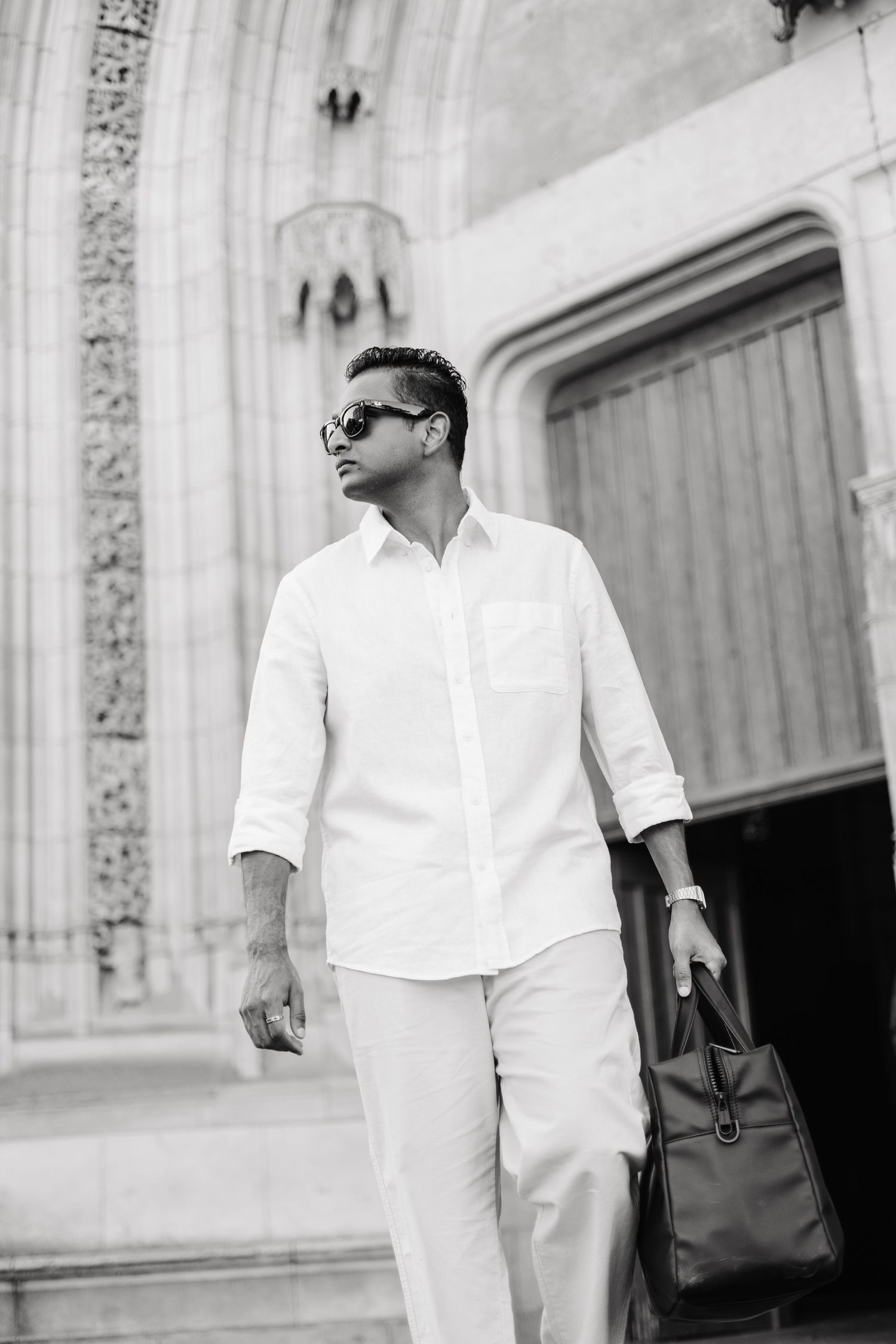 Black and white photo of a stylish man in white shirt and pants, wearing sunglasses, holding a leather bag outside a building.