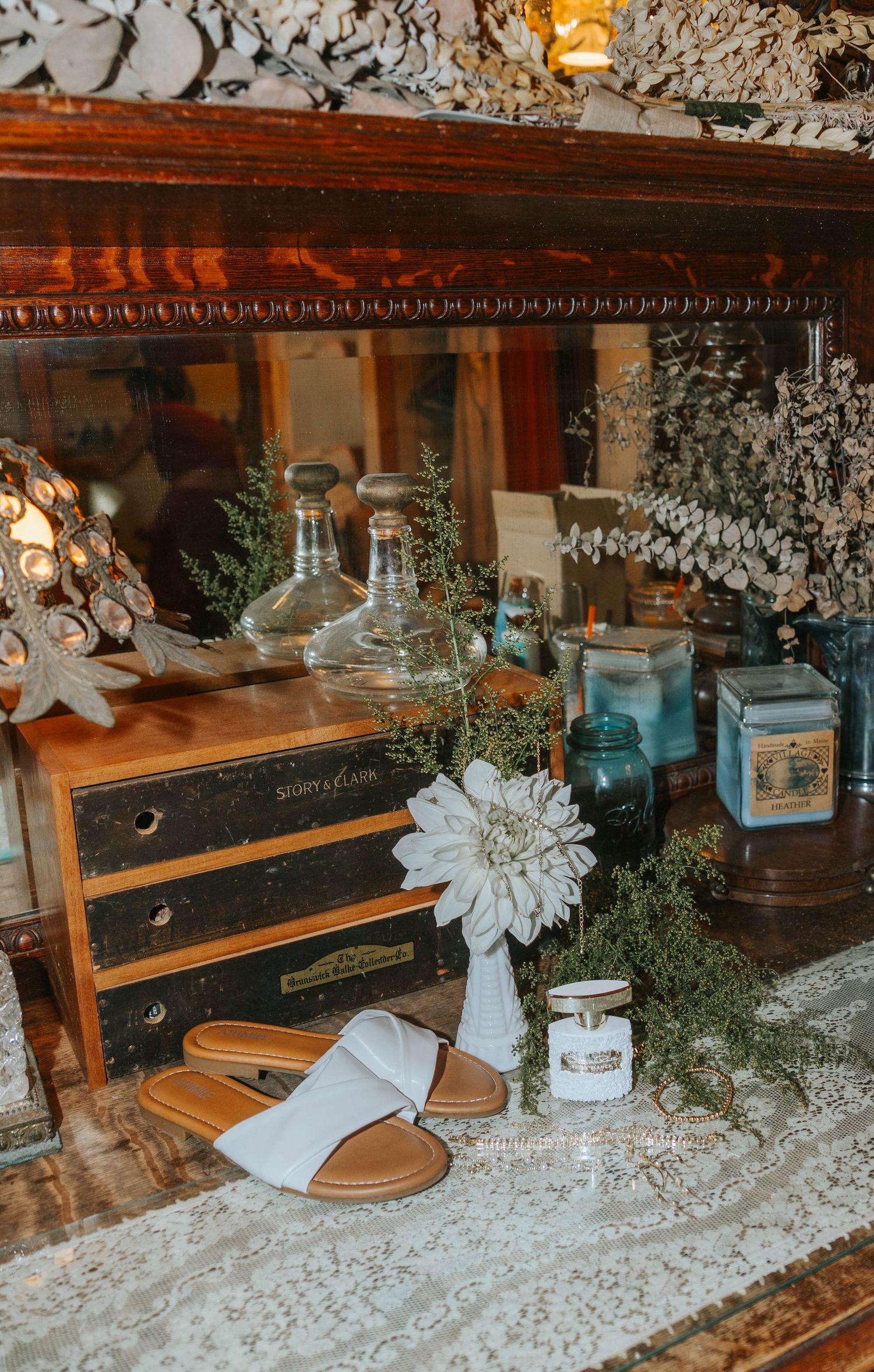 Vintage display with tan sandals, white dahlia, glass decanters, and antique drawers on lace-covered surface.