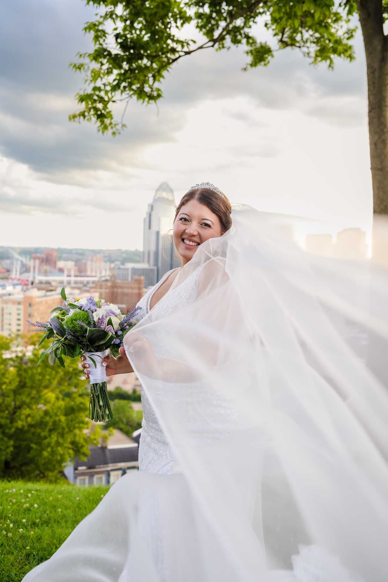 Smiling bride in white gown and flowing veil holds bouquet outdoors with city skyline backdrop at the view wedding venue