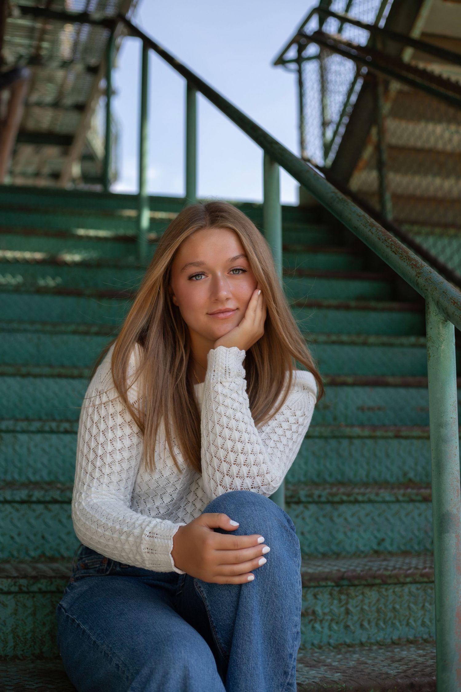 Young woman in white sweater and jeans sitting on green metal stairs, resting chin on hand.