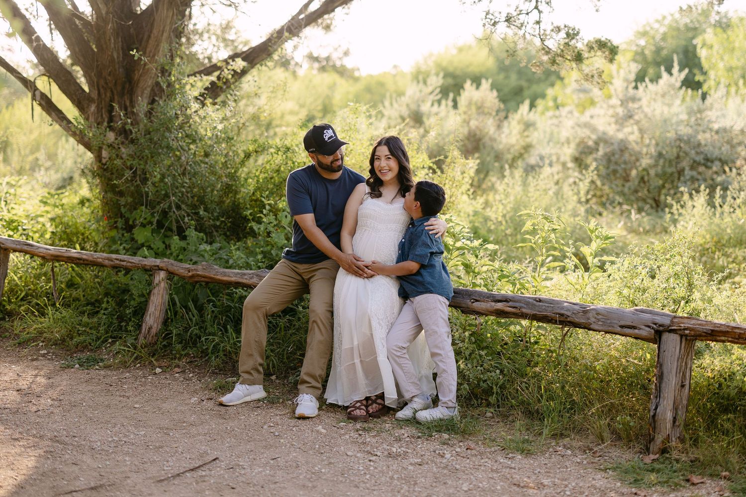 A family of three poses outdoors on a rustic wooden fence, with a pregnant woman in a white dress surrounded by lush greenery.