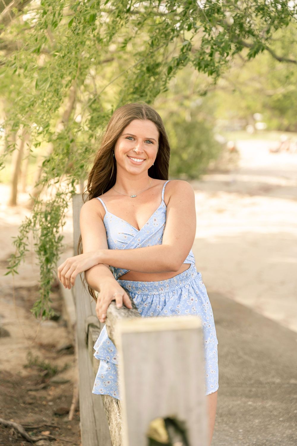 Portrait photo of a brunette woman along the beach leaning on a fence with a willow tree behind her